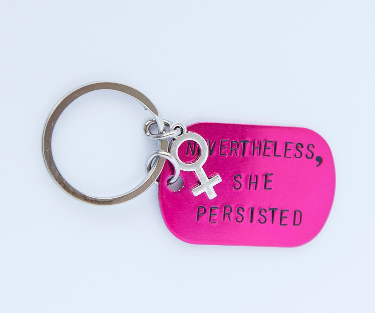 A pink feminist keychain with the phrase 'Nevertheless She Persisted' hand-stamped on a lightweight aluminum tag, attached to a durable stainless steel ring.